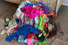 Load image into Gallery viewer, Pile of colorful bikinis and swimwear fabric on a textured surface next to a decorative wooden cabinet.
